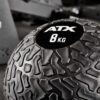 Close-up of a black ATX® Slam Ball labeled 8 KG with a textured surface, set against a blurred gym background.