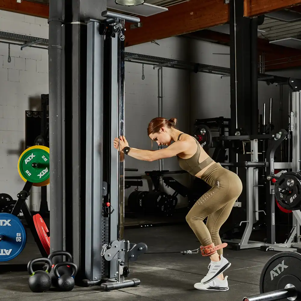 A woman exercises at a gym, using a single cable machine. She wears a beige workout outfit and ankle straps, performing a leg movement. Gym equipment, including weights and kettlebells, is visible in the background.