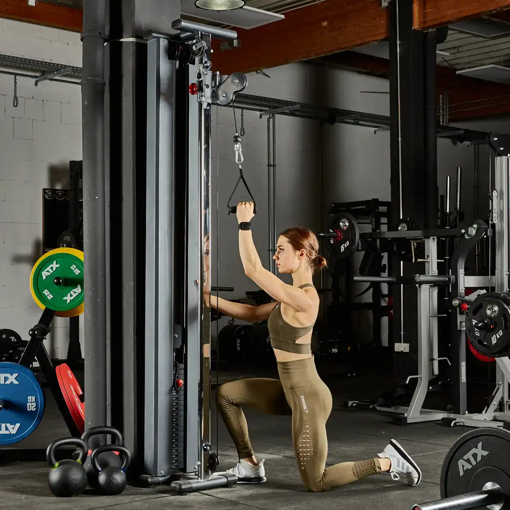 A woman kneels next to a wall-mounted cable machine in a gym, preparing to exercise. She wears a brown athletic outfit. The gym's backdrop is filled with various equipment, including weights and kettlebells.
