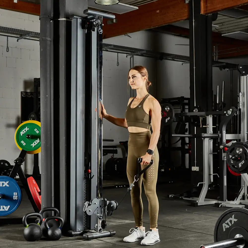 A woman in athletic attire stands in a gym holding the handle of a wall-mounted cable machine. She's wearing a brown sports bra and leggings with white sneakers. The gym is equipped with weights and kettlebells in the background, setting the scene for her intense workout session.