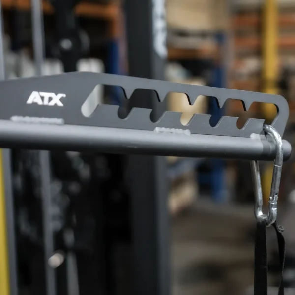 Close-up of the ATX Multi X Long Bar 90cm, a gray metal arm with black straps connected by a carabiner, set in front of blurred gym equipment and shelves.
