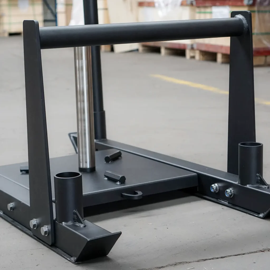 The Power Maxx Gym Weight Sled, featuring a vertical pole for weight plates and angled handles ideal for pushing or pulling, is shown resting on a concrete floor. This makes it perfect for strength training and fitness exercises. Wooden pallets in the background enhance the rugged gym atmosphere.