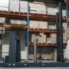 The Power Maxx Gym Weight Sled, featuring a metal pole for weights, sits on the concrete floor of a warehouse. Behind it, shelves filled with various boxes and equipment provide an industrial backdrop.