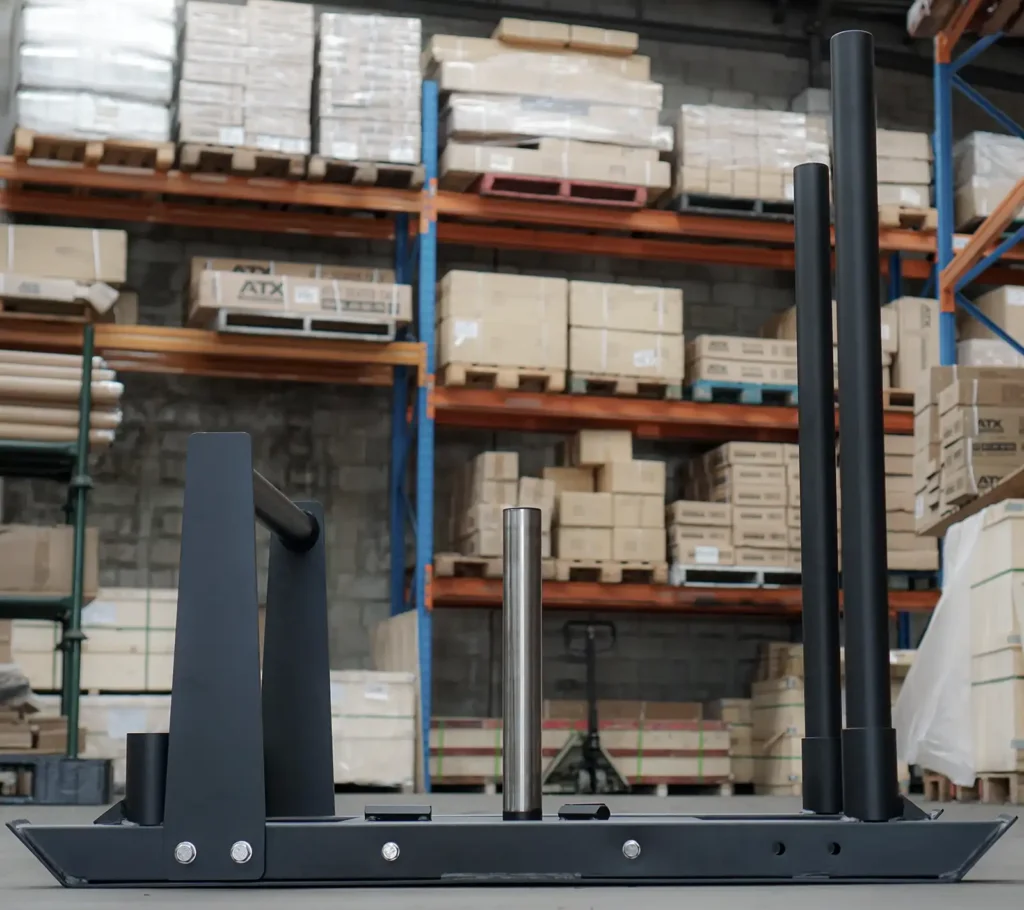 The Power Maxx Gym Weight Sled, featuring a metal pole for weights, sits on the concrete floor of a warehouse. Behind it, shelves filled with various boxes and equipment provide an industrial backdrop.