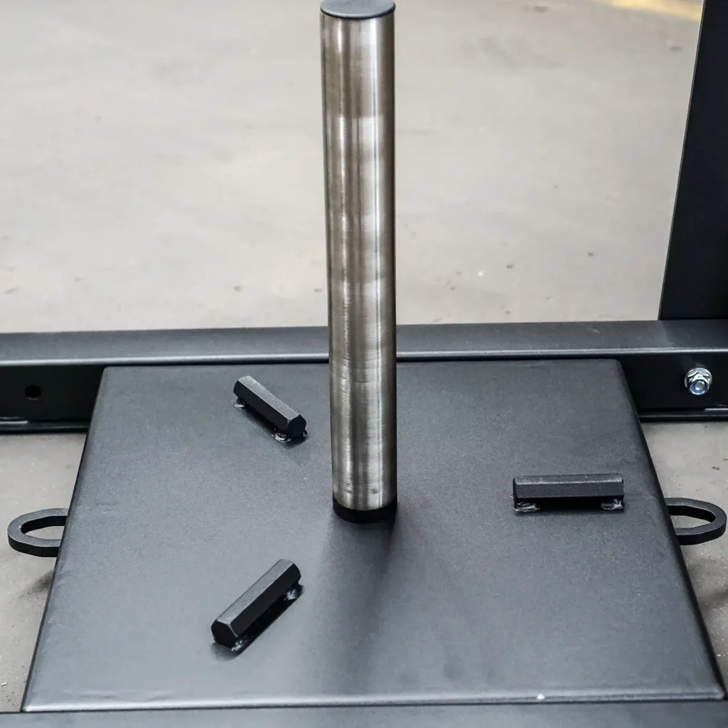 Close-up of a Power Maxx Gym Weight Sled on a smooth, neutral-toned gym floor. The sled consists of a vertical steel rod mounted on a square base, complete with three small brackets for securing weights.