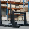 A Power Maxx Gym Weight Sled with vertical handles rests on the warehouse floor, where shelves brimming with cardboard boxes are visible in the background.