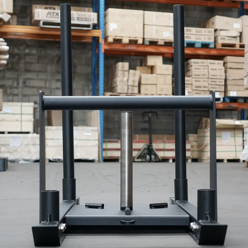 A Power Maxx Gym Weight Sled with two vertical handles is positioned in a warehouse setting. Cardboard boxes are stacked on shelves in the background, adding to the busy, industrial feel of the space. The concrete floor and lighting enhance the rugged ambiance of this pull-push sled environment.