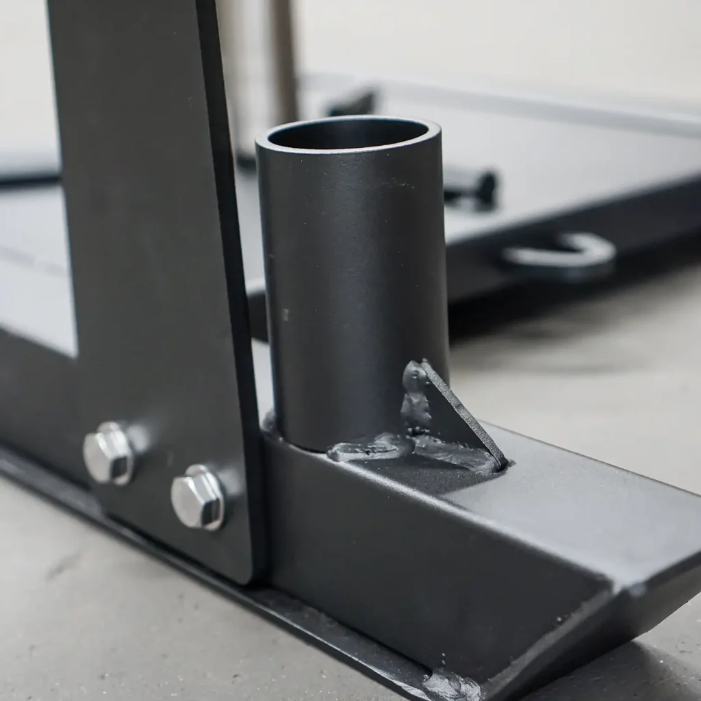 Close-up of the Power Maxx Gym Weight Sled, a black metal structure with bolts and a cylindrical component. The smooth, industrial surface suggests it's part of mechanical or construction equipment.