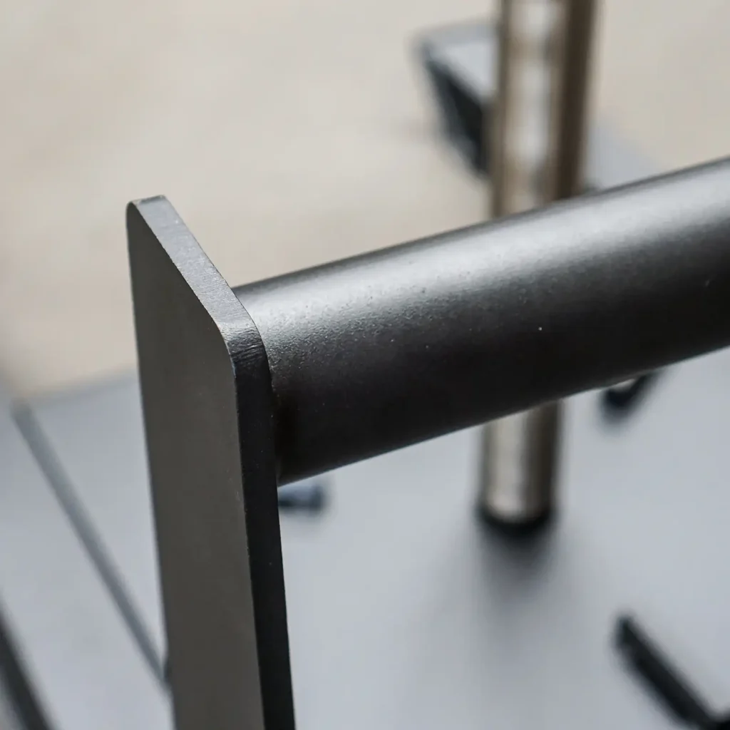 Close-up of the Power Maxx Gym Weight Sled, highlighting its horizontal cylindrical rod and vertical flat plates with a smooth, matte finish. The blurred background accentuates the sled's industrial design elements.