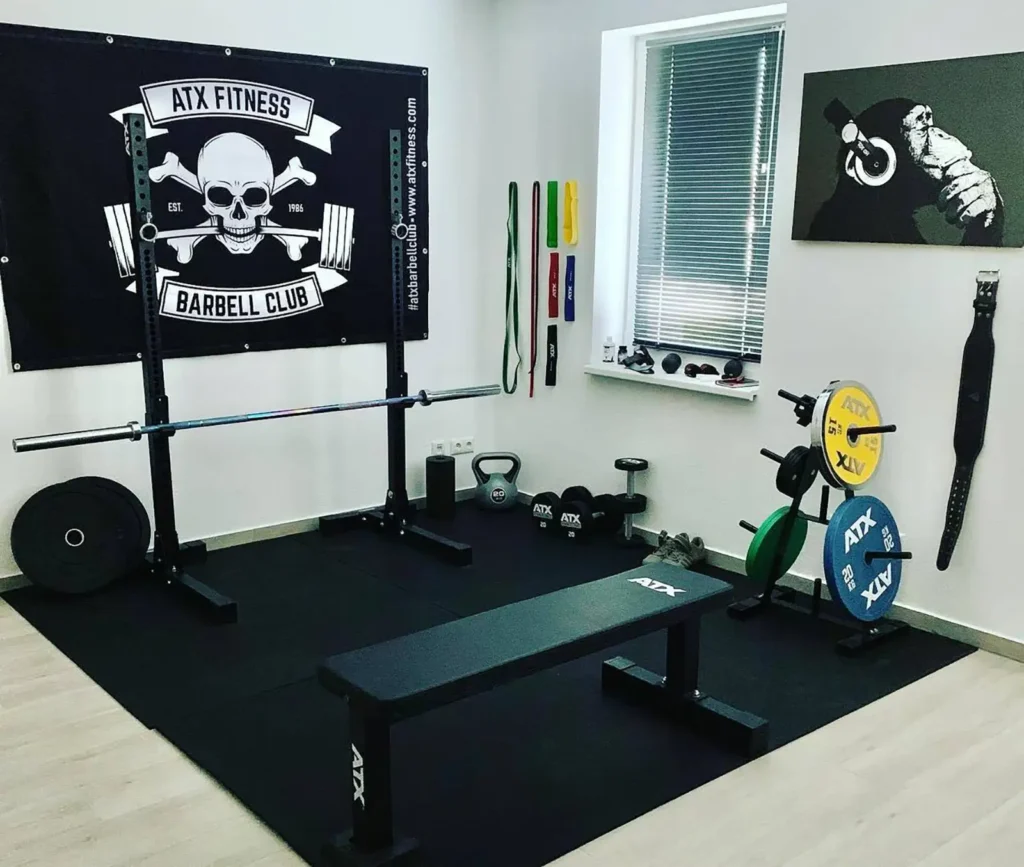 A home gym featuring a flat weight lifting bench, barbell, and colorful weight plates. A banner on the wall reads ATX Fitness Barbell Club. Various resistance bands and kettlebells are neatly arranged, alongside a motivational poster.