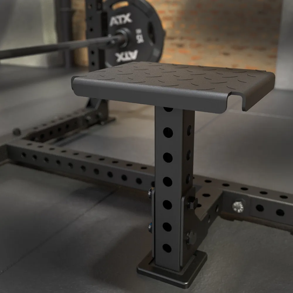 A close-up of the ATX® Seal Row Bench, black and adjustable, mounted to a power rack in a gym with a barbell, weight plate, partial floor, and brick wall visible in the background.