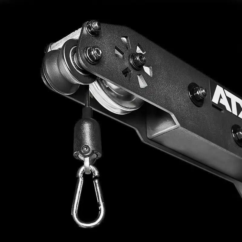 Close-up of a metal pulley with hook and cable, set against a black background. The mechanism, part of the ATX® Triplex Weight Stack Gym, features bolts and cut-out details typical of gym equipment.
