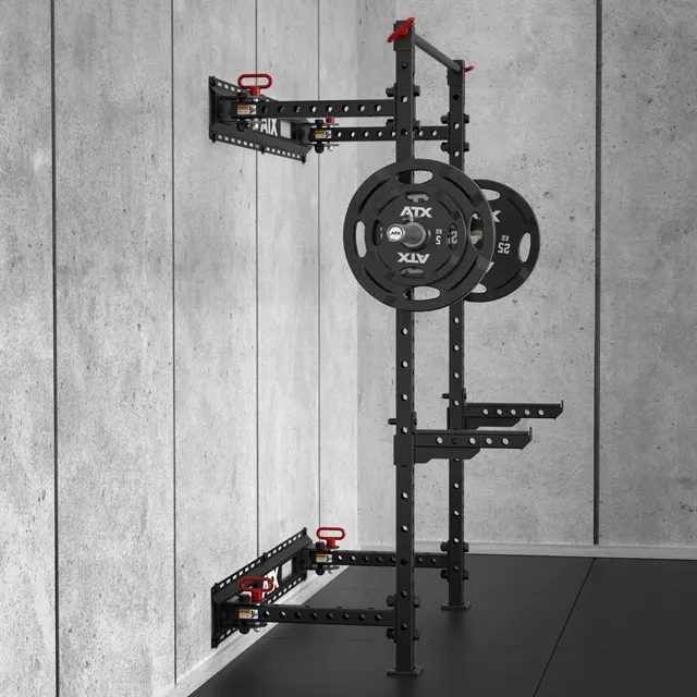 The ATX® Folding Squat Rack 500, a black wall-mounted rack with red adjustment knobs and multiple height holes, is set up with weights against a gray concrete wall in a gym with black flooring.