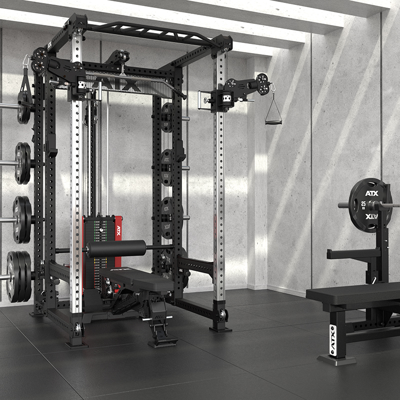 A modern home gym showcases the ATX® Power Cage with Cables, weight plates, a cable machine, and a bench press on black mats in a concrete-walled room with exposed beams.