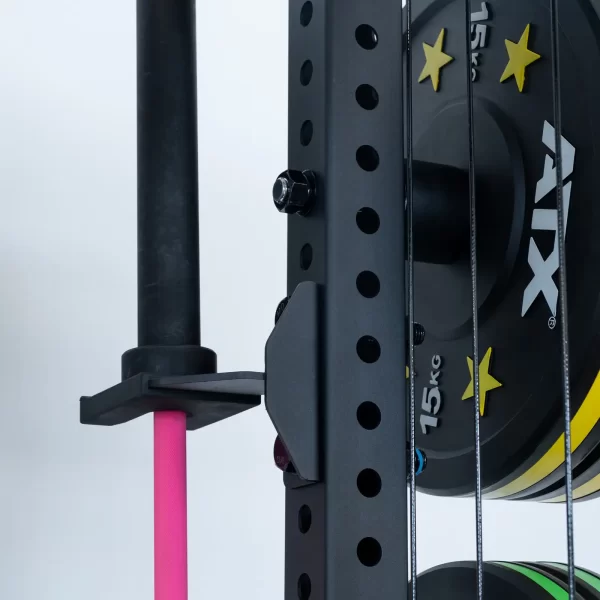 Close-up of the ATX® Power Cage with cables, featuring a pink barbell and weight plates. The rack has holes and a black metal holder. A 15kg ATX plate is prominently displayed, with more plates stacked behind the barbell.