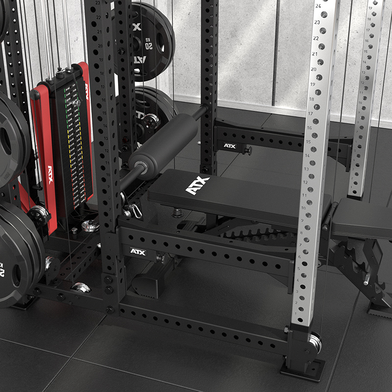 Close-up of an ATX® Power Cage with Cables, equipped with weight plates, a padded bench, and leg roller attachment, placed on tiled flooring in a modern fitness room with concrete walls.