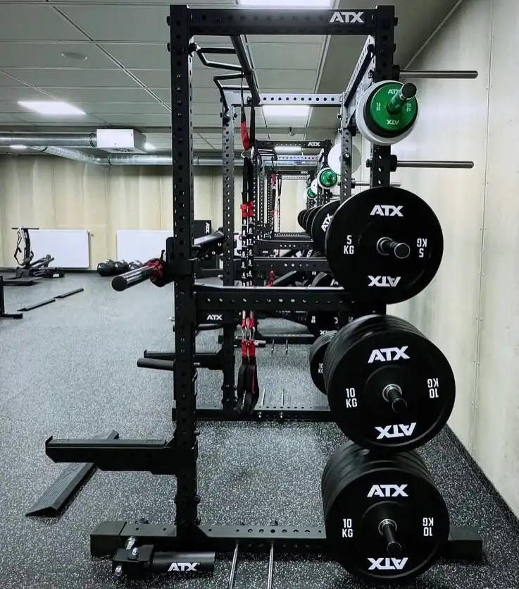 A Commercial Power Rack 780 in a gym holds various black ATX weight plates and barbells; the gym features gray flooring, light walls, and other exercise equipment in the background.