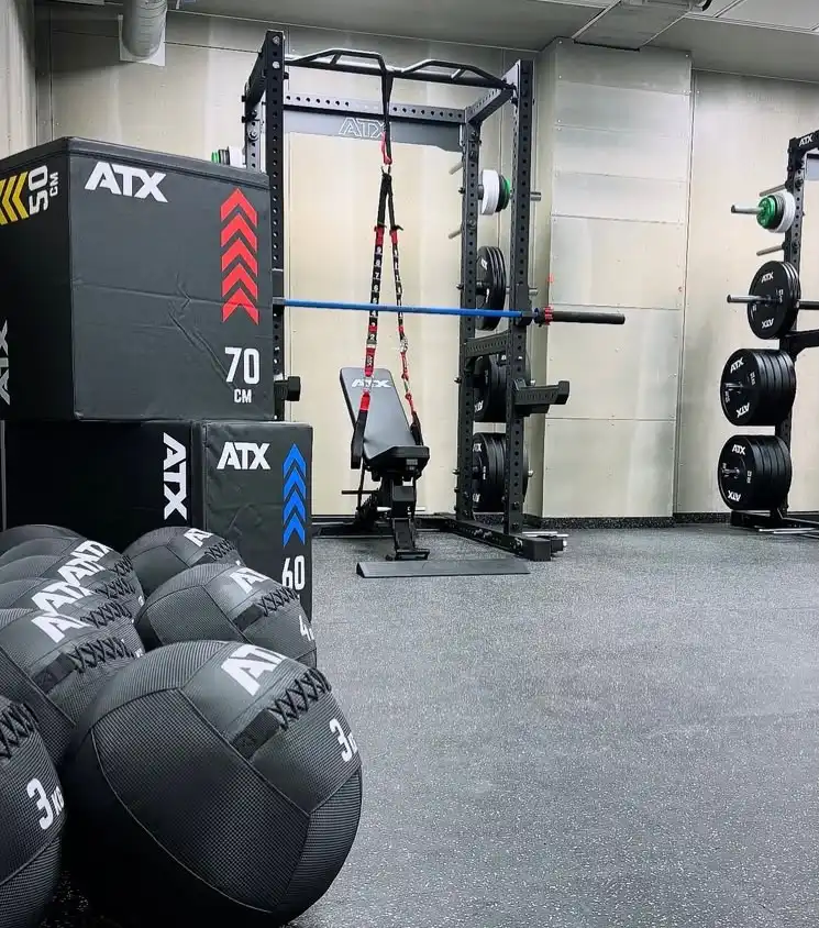 A gym featuring the Commercial Power Rack 780, a bench, weight plates on racks, stacked plyometric boxes, and several medicine balls. The space is clean with minimal decor and black rubber flooring.