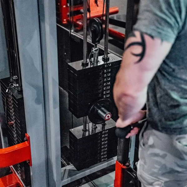 In a gym, someone uses a resistance machine, their arm and upper body partially visible. A tribal tattoo graces their upper arm. They expertly adjust the GymPin 2 Original, wearing a gray shirt as they push their limits.