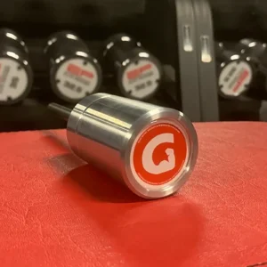 A close-up of a metallic GymPin 2 Original handle, showcasing an orange logo with a stylized G on the end. The handle is positioned on a red surface, with blurred weights in the background.