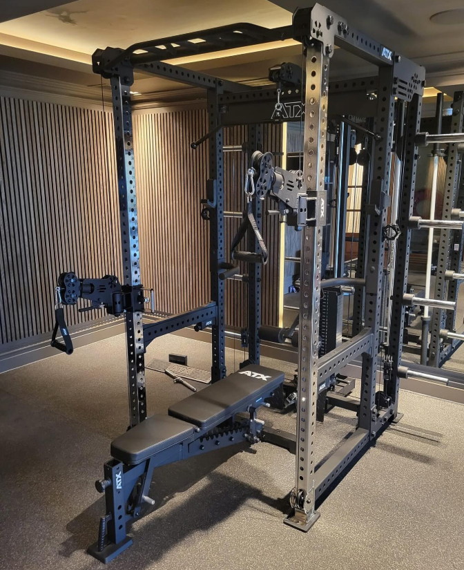 An ATX® Power Cage with Cables, featuring various attachments and a bench, is set up in a well-lit carpeted room with wooden wall panels.