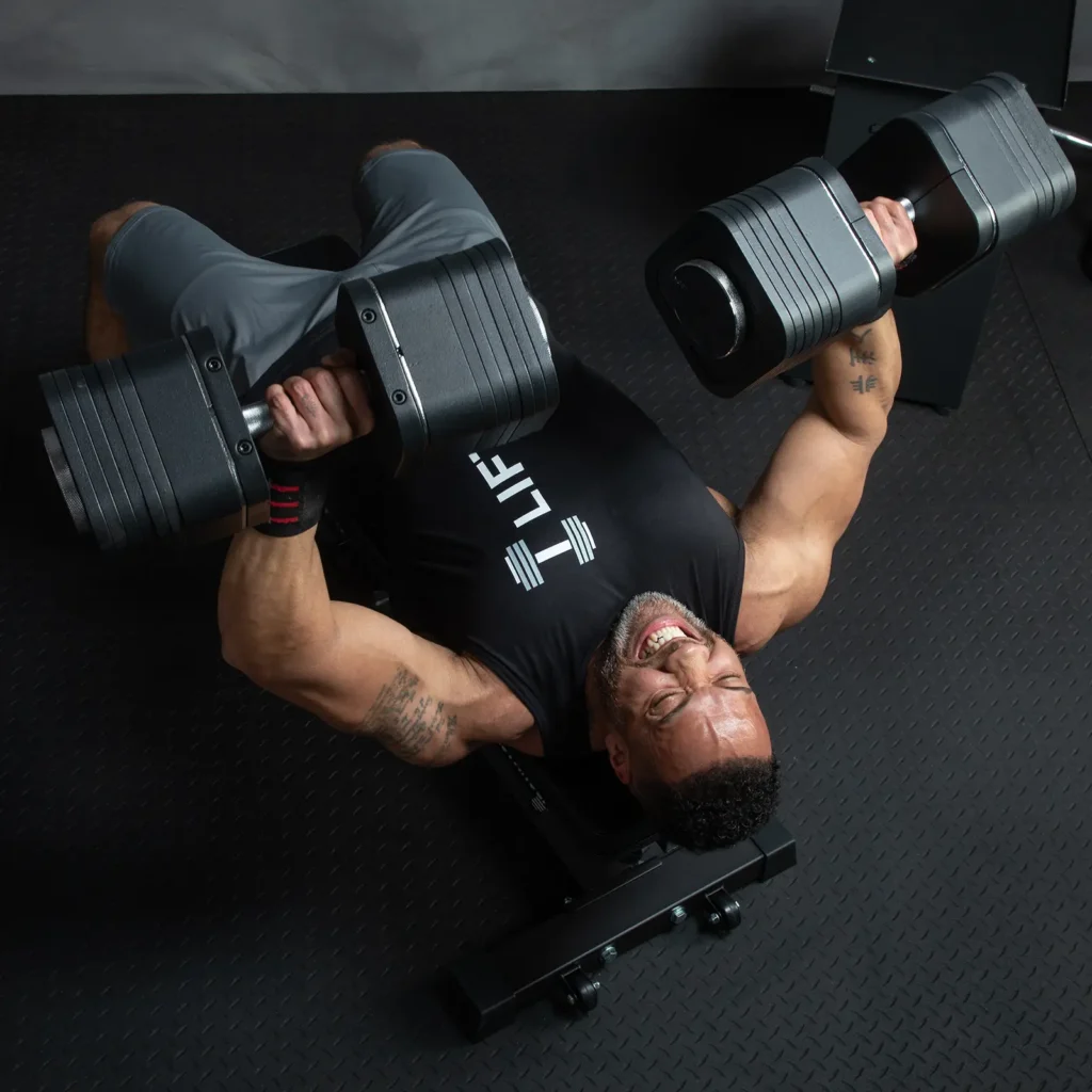 A person with a muscular build is lying on a bench, lifting large Ironmaster adjustable dumbbells. They are wearing a black tank top and gray shorts, completely focused on the exercise in a gym setting with a dark floor.