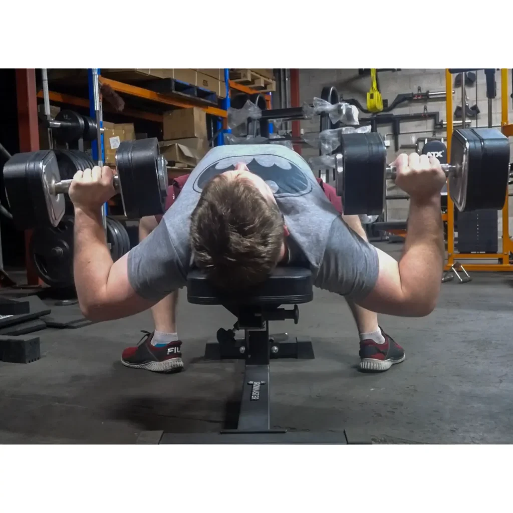 A person is lying on a bench in a gym, performing a bench press with Ironmaster adjustable dumbbells. They are wearing a gray T-shirt and sneakers. The background showcases gym equipment and shelves with boxes, adding to the focused atmosphere of their workout.