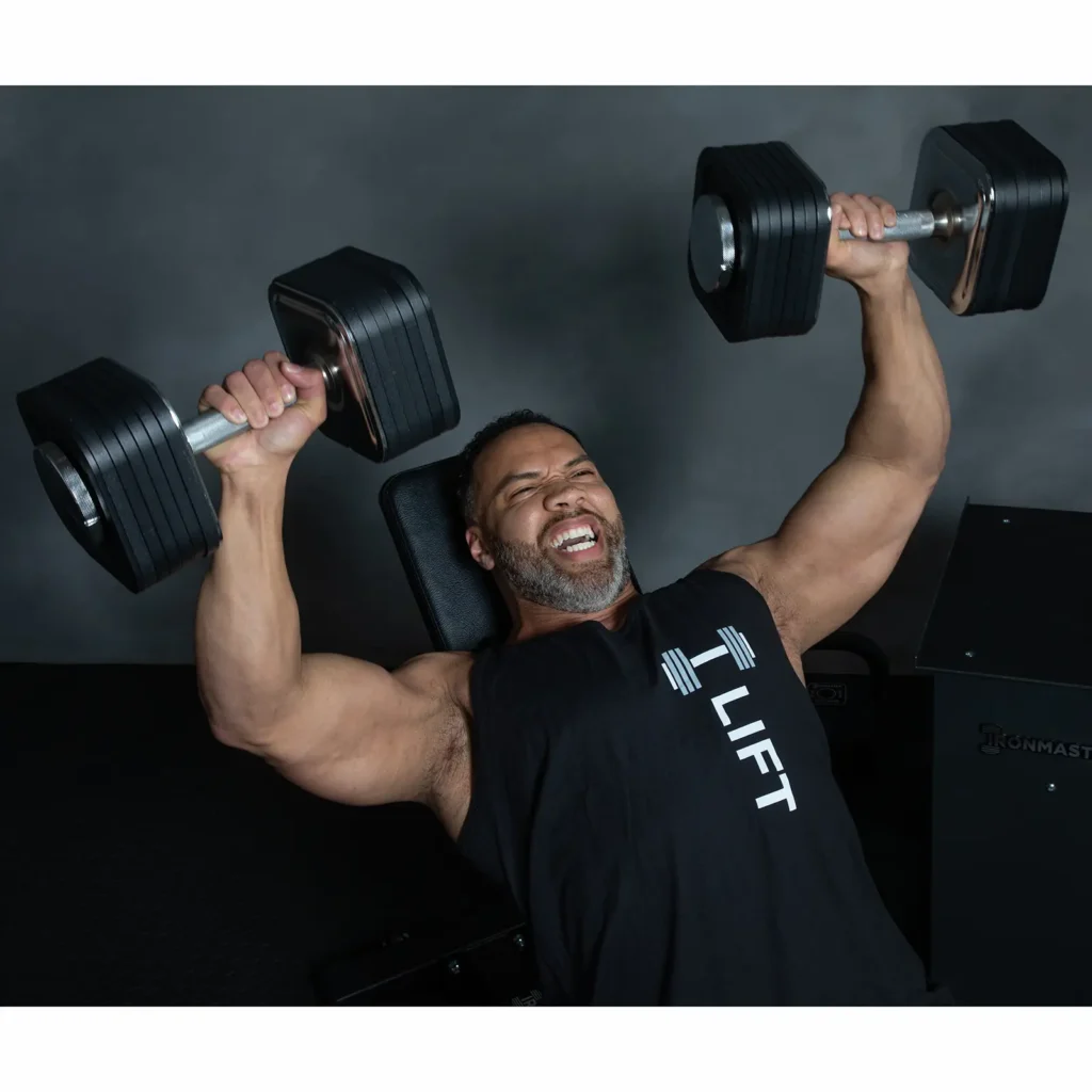 A person with a beard is performing a chest press using Ironmaster adjustable dumbbells on a bench. They wear a black sleeveless shirt emblazoned with LIFT. The dark background accentuates their unwavering effort and focus.