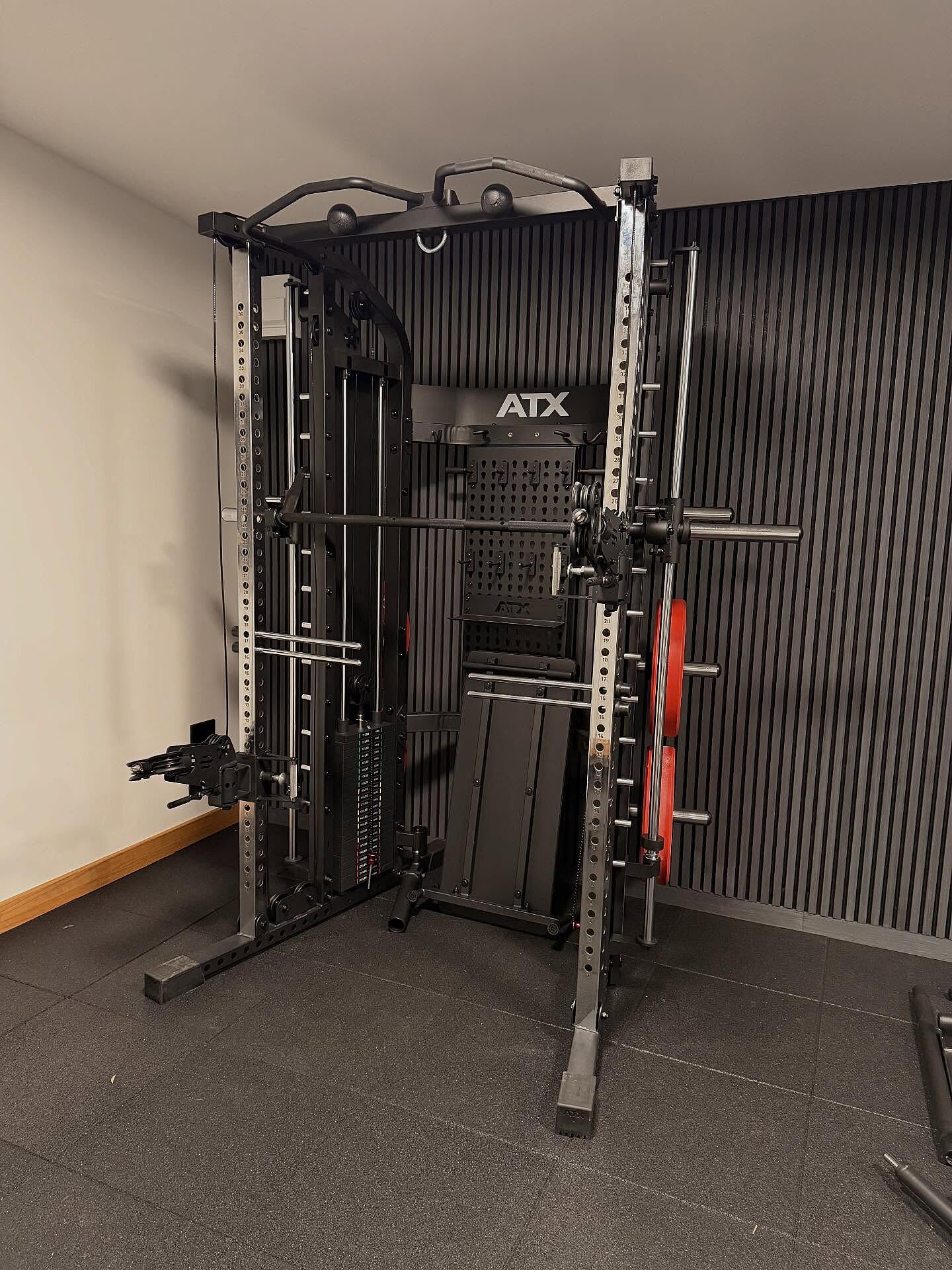 A home gym setup featuring the ATX® Monster 2.0 All In One Trainer in black, equipped with a pull-up bar, weight plates, and a bench, set against a grooved black wall on a rubber floor.