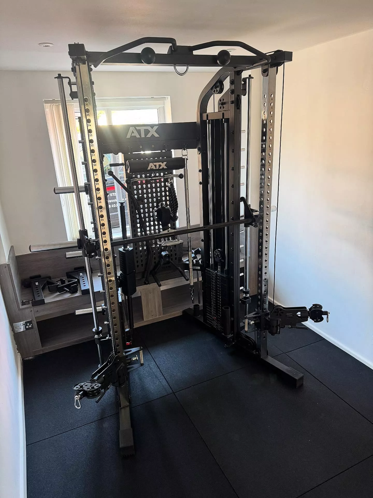 A home gym features an ATX® Monster 2.0 All In One Trainer with barbell, cable pulleys, attachments, and weight storage on black rubber flooring in a bright, minimalist room.