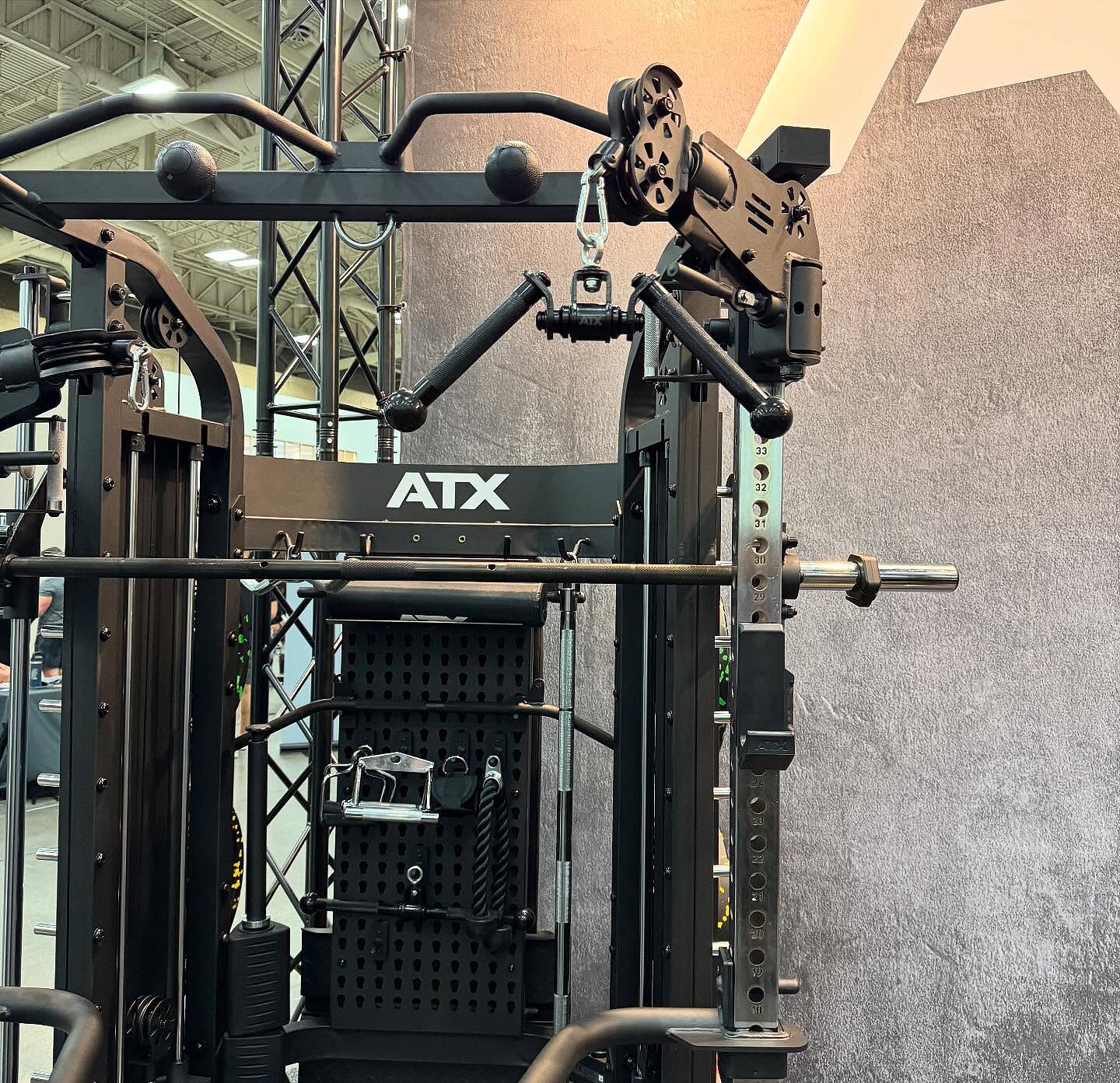 The ATX® Monster 2.0 All In One Trainer, featuring multiple attachments, a pull-up bar, adjustable cables, weight plates, and a barbell, is showcased against a gray wall in a fitness facility.