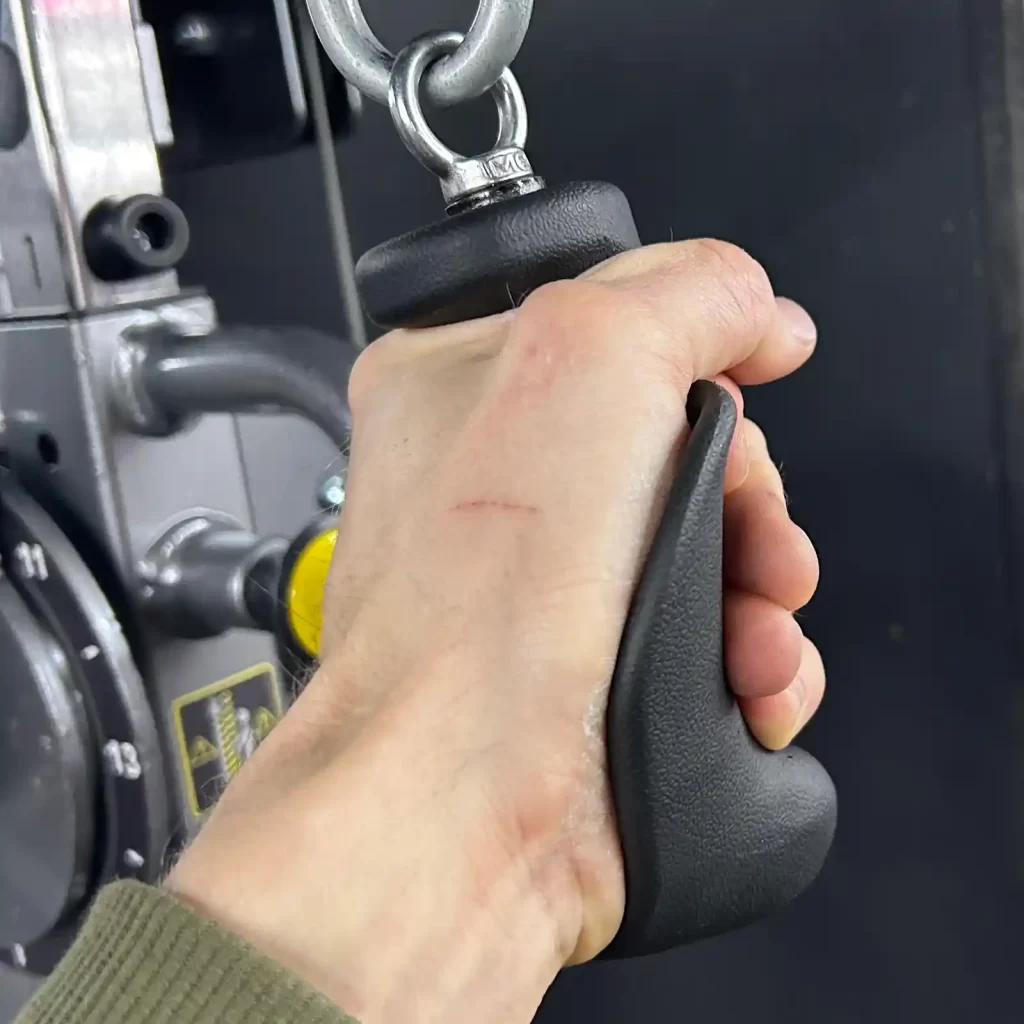 A close-up of a hand gripping the black Spud Inc Gorilla Fingers gym cable attachment handle, connected to a cable machine with a metal carabiner. The hand, showing a small scratch, is wearing a green sleeve.
