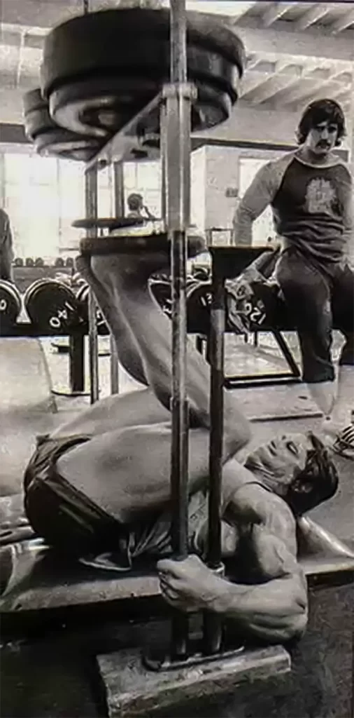 Someone is using the ATX® Vertical Leg Press, pushing weighted plates with their legs while on their back. In the background, a person with a mustache and gym attire observes, amidst the lively atmosphere of a bustling gym.
