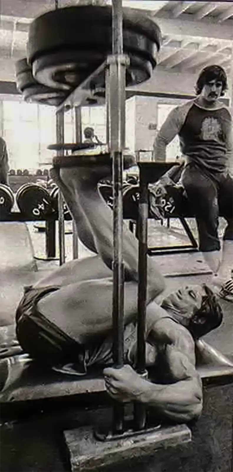 Someone is using the ATX® Vertical Leg Press, pushing weighted plates with their legs while on their back. In the background, a person with a mustache and gym attire observes, amidst the lively atmosphere of a bustling gym.