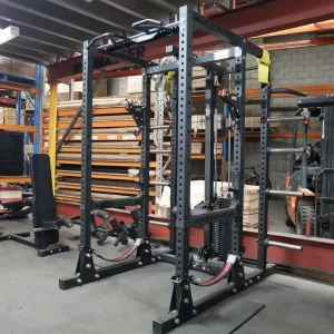 A black metal power rack set up in a warehouse gym, equipped with various attachments and pulleys. The background shows industrial shelves with wooden planks and a forklift. The space appears to be multifunctional for storage and exercise.