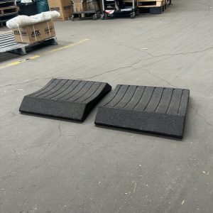 Two dark, rectangular ramps placed on a concrete floor in a warehouse. The ramps have grooved surfaces and are positioned side by side. In the background, there are boxes and pallets on shelving units.