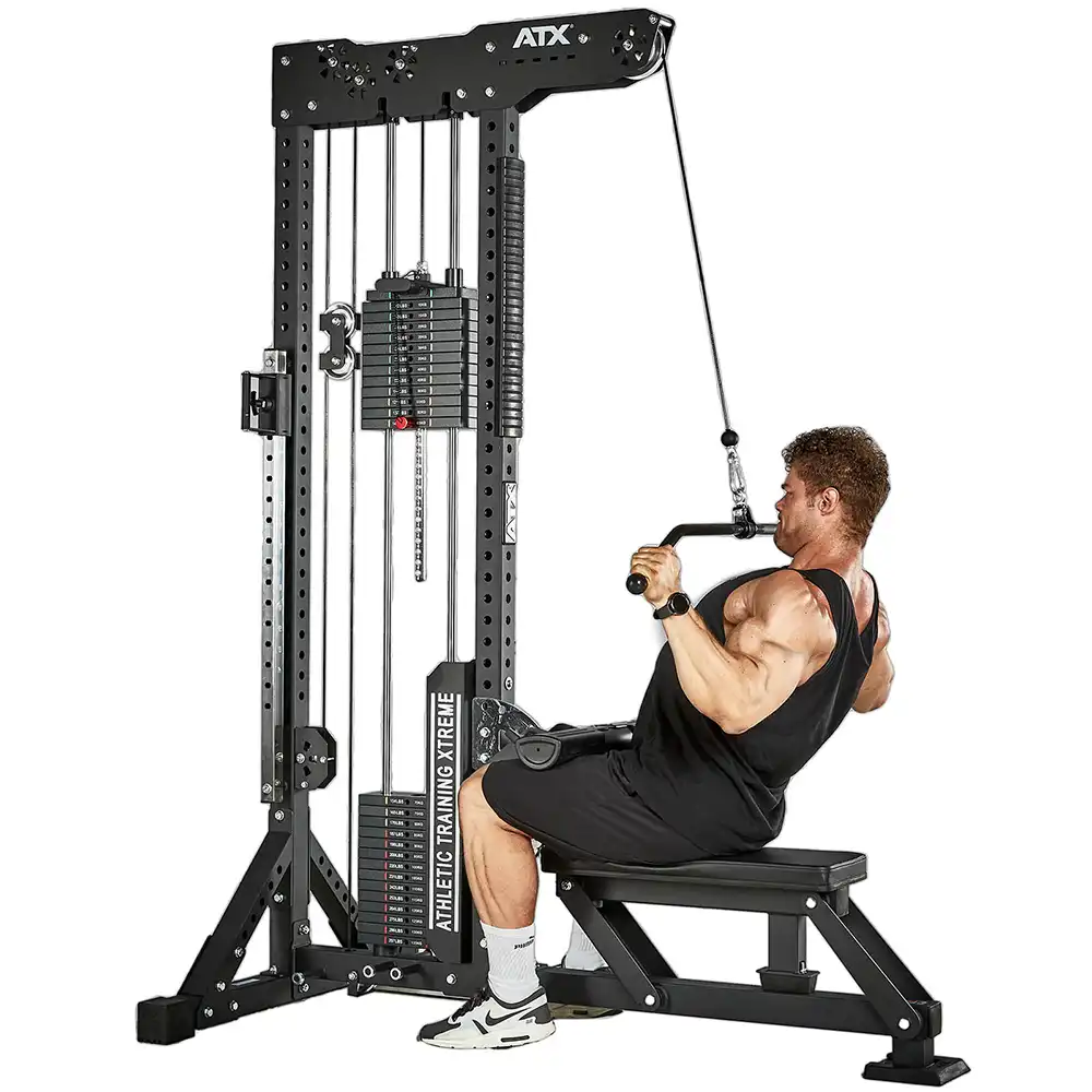 A person in a black tank top and shorts is using a cable machine for lat pulldowns, sitting on a bench. The machine is labeled ATX and has adjustable weight stacks.