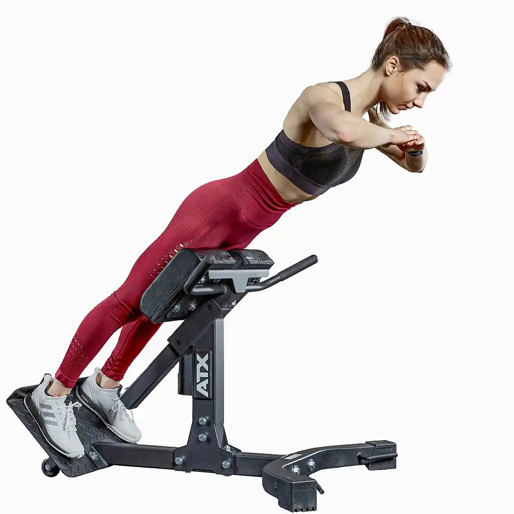 A woman in athletic wear performs a back extension exercise on a fitness bench. She is positioned with her body parallel to the ground, arms crossed over her chest, and legs secured under pads. Her athletic wear is a matching red set.