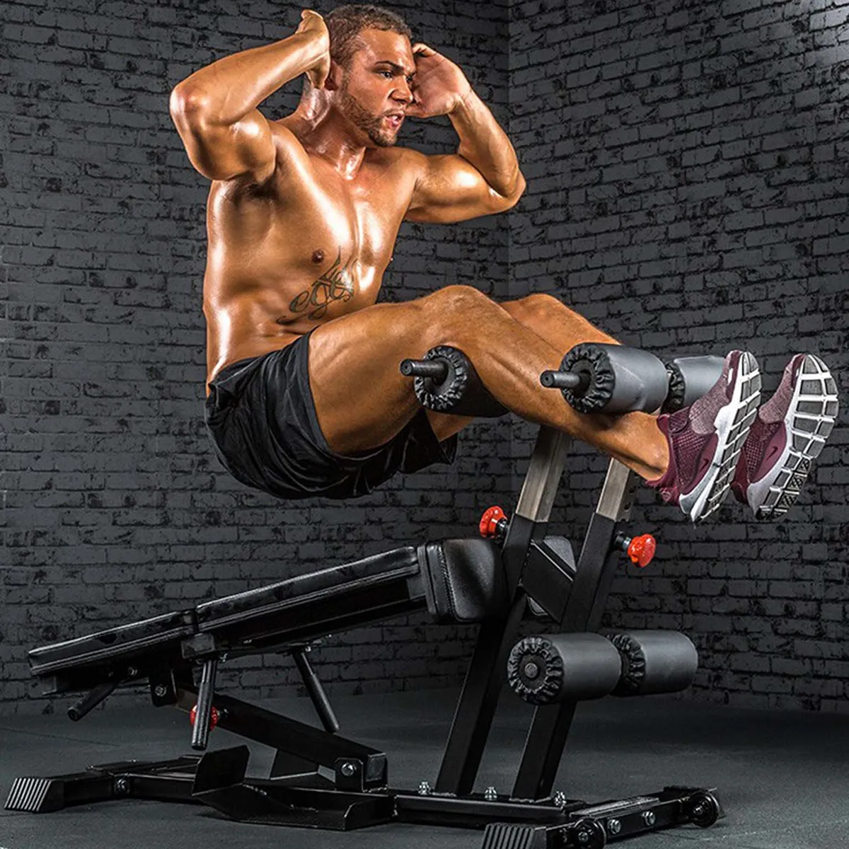 A muscular, shirtless man in black shorts and maroon running shoes performs sit-ups on the ATX Torso Trainer, focusing intensely in a gym with dark brick walls.