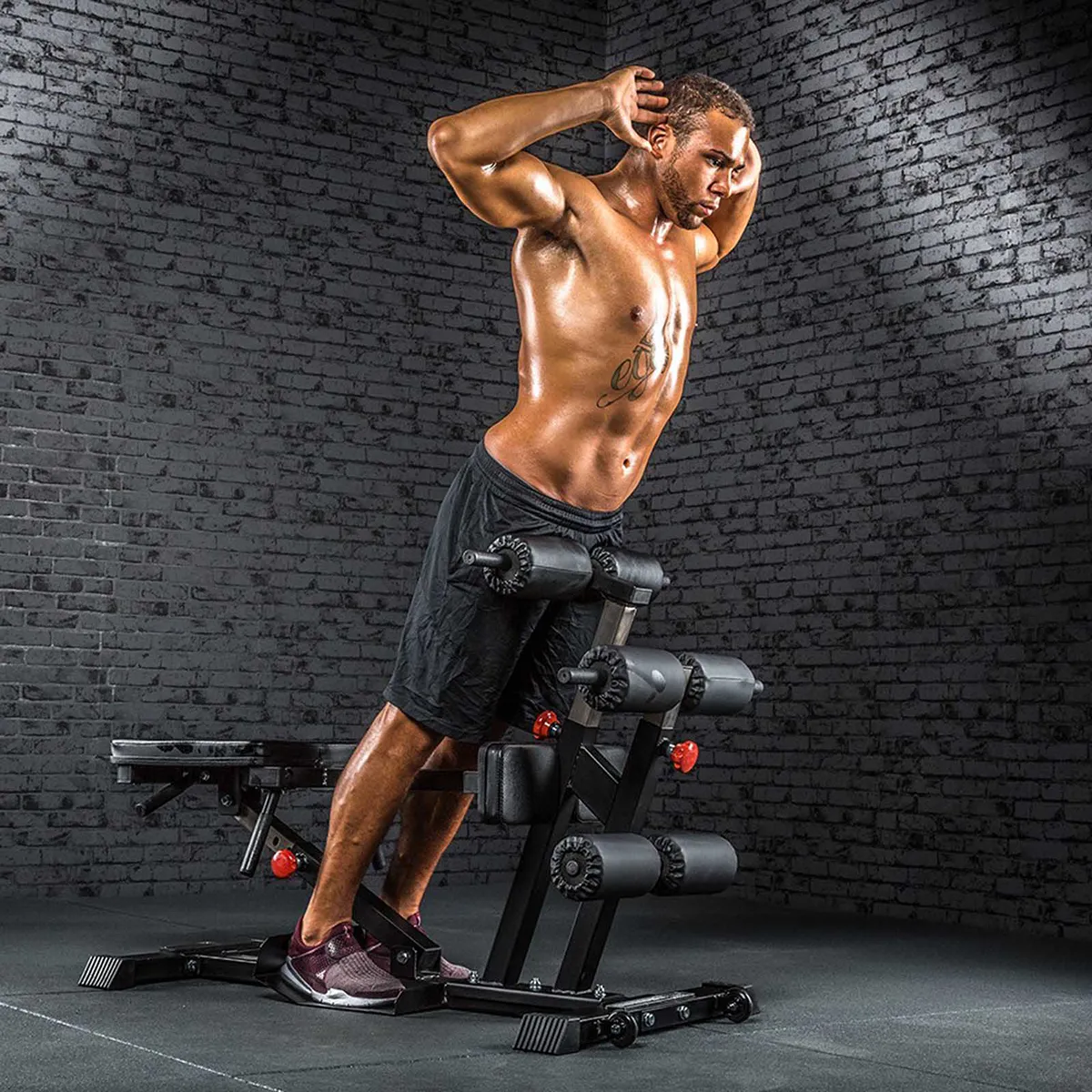 A muscular man in black shorts and sneakers performs a back extension on the ATX Torso Trainer next to an ab machine in a gym with dark brick walls, hands behind his head and upper body lifted.