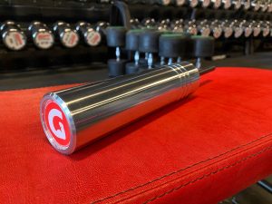 A metallic cylindrical barbell handle with a red and white end cap logo rests on a red bench. Rows of dumbbells with labels are blurred in the background, set in a gym environment.