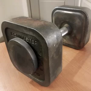 A close-up of a single dumbbell with a square, metallic design on a wooden surface. The label reads IRON MASTER, and it has a textured grip handle. The weight marking indicates 120 LB.