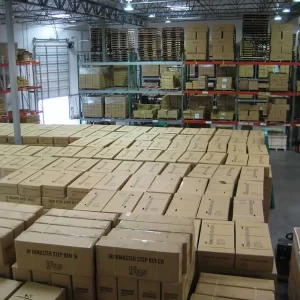 Warehouse interior filled with stacks of cardboard boxes, neatly arranged in rows. Shelving units in the background hold more boxes and pallets. A large overhead door is partly visible on the left. The space is well-lit.