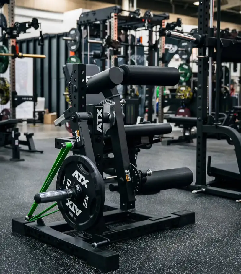 A glute ham developer with black padding and weights is positioned among various strength training equipment, including the ATX® Seated Leg Curl Extension Machine and multiple power racks in a well-equipped gym.