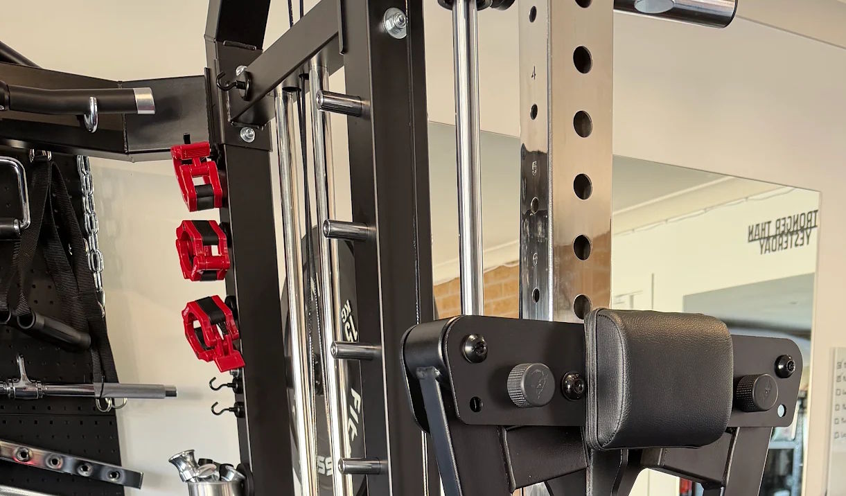 Close-up of gym equipment, featuring a black padded arm support and steel bars with adjustable holes designed to accommodate the ATX® Lateral Raise Attachment, as well as red attachments and a mirror reflecting a PROCORE TEAM sign in the background.