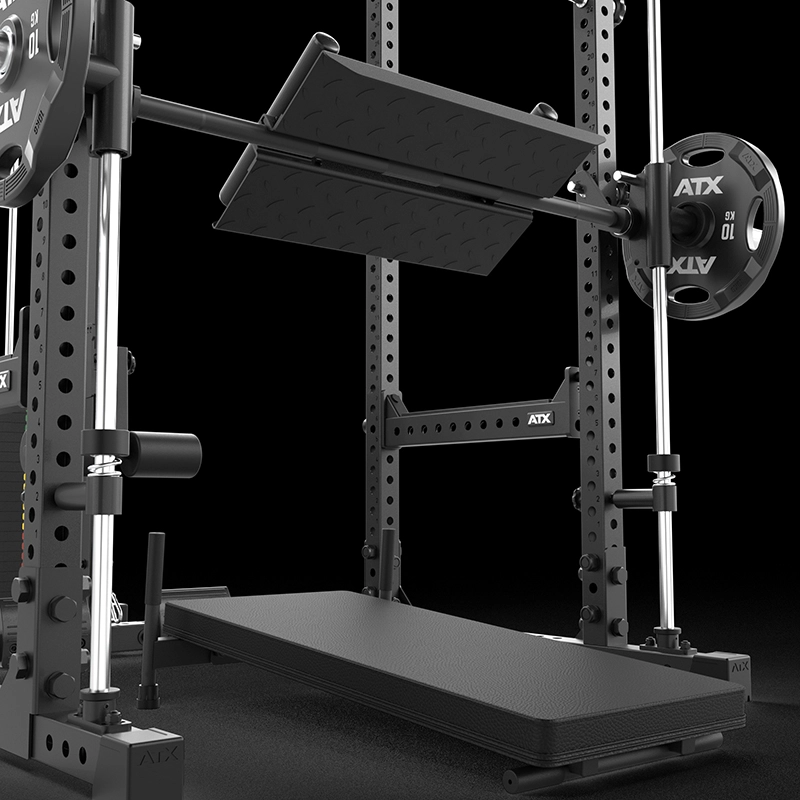Close-up of a black power rack with a loaded barbell and spotter arms. A padded bench and angled footplate are positioned underneath, ready for a bench press or squat exercise. The equipment is branded ATX.