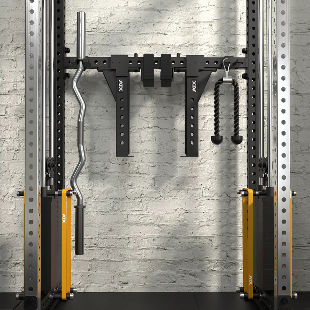 A gym equipment rack mounted on a white brick wall holds a curl bar, two black angled attachments, and a double tricep rope, framed by metal squat rack posts with yellow safety catches.