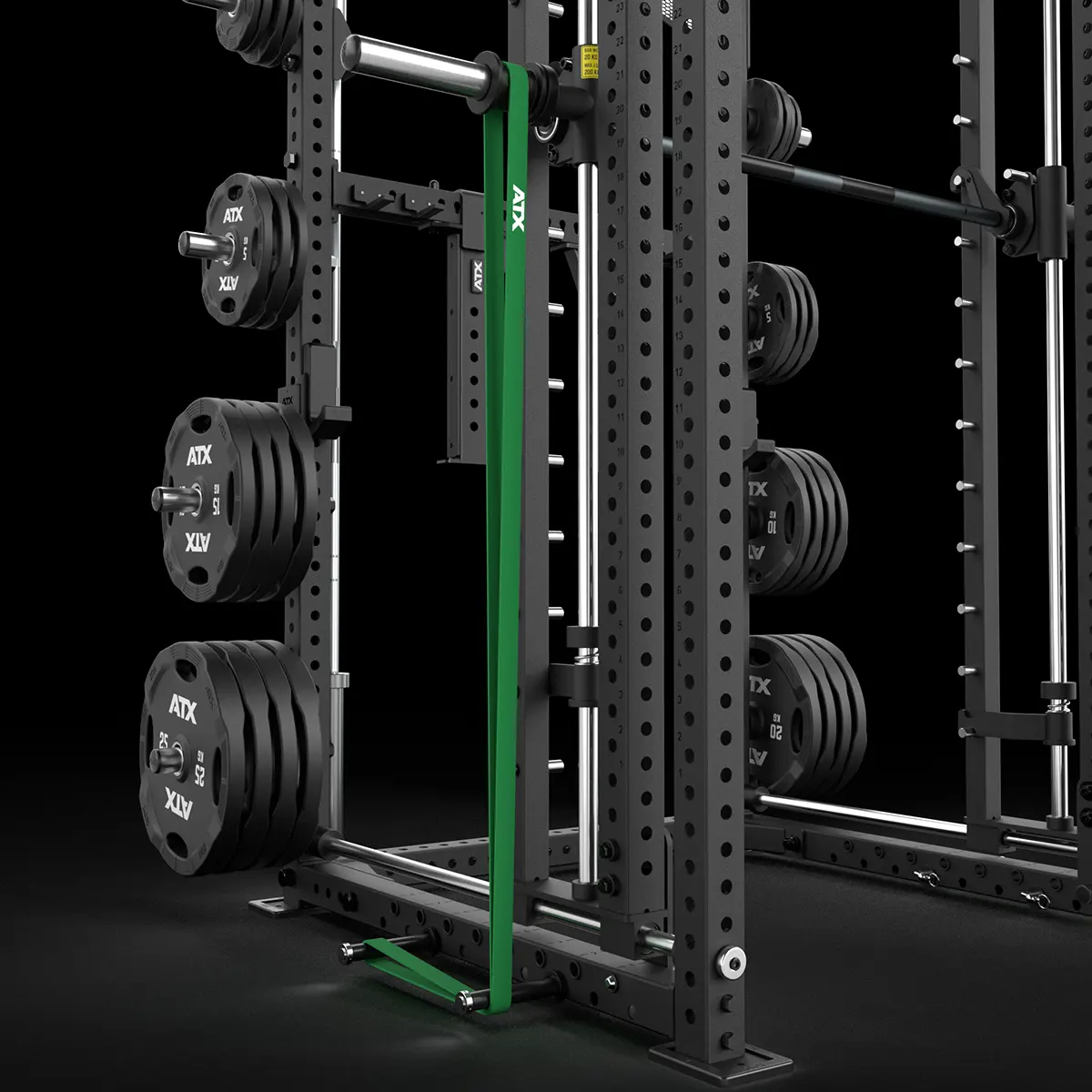 A black power rack with several weight plates stored on its side and a green vertical lever arm attachment. The setup is positioned on a black floor against a dark background.