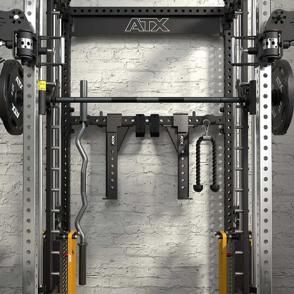 A gym power rack with an ATX logo, holding a barbell, weight plates, curl bar, rope attachment, and padded handles, set against a white brick wall. Various attachments are organized on the rack.