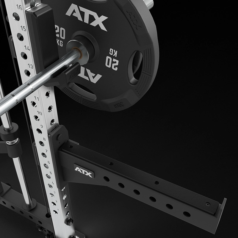 A close-up of the ATX® Squat Rack with Cables (Copy), showcasing a loaded barbell with a 20 kg ATX weight plate, adjustable safety arms, and metallic support columns with numbered holes, set against a black background.